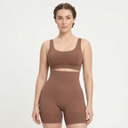 A woman modeling a matching brown activewear set consisting of a scoop-neck sports bra and high-waisted compression shorts, shot against a plain light gray background.