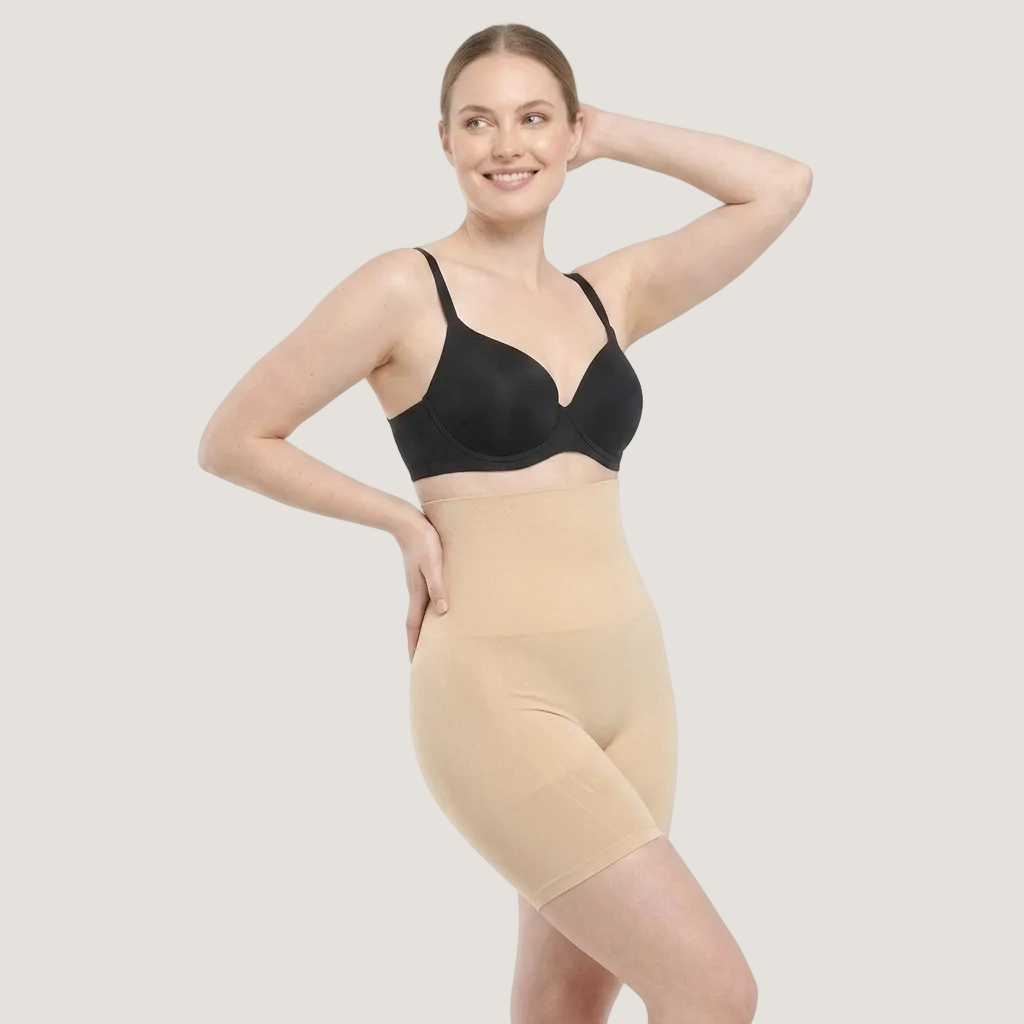 A woman modeling a beige high-waisted compression shapewear brief over a black bra, posed against a clean white studio background.